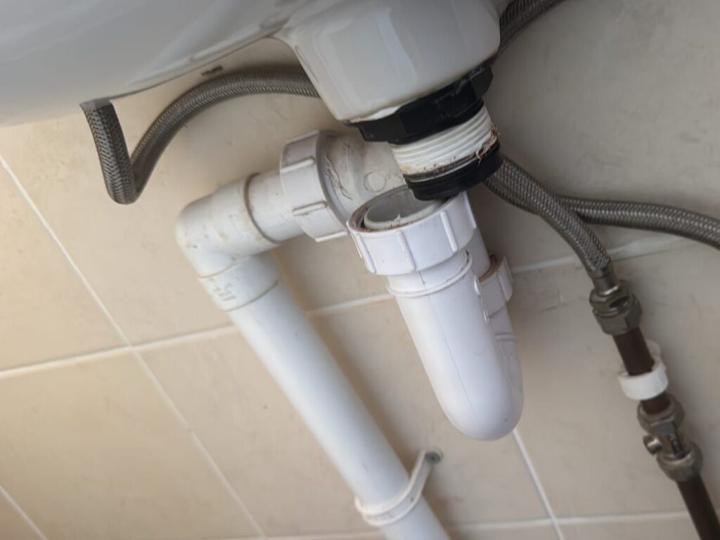New white bottle trap and waste pipework under a basin, with braided supply hoses.